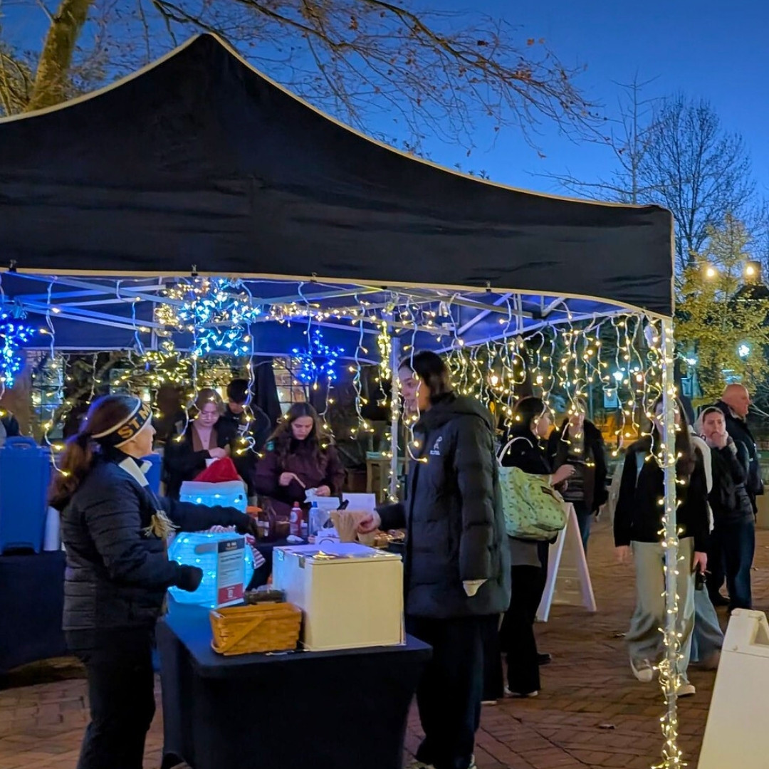 People gather under a black canopy decorated with string lights at an outdoor evening event, with tables set up for food or drink service.
