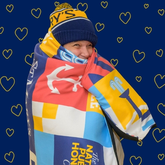 A person bundled in a colorful patterned blanket and blue winter hat smiles against a dark blue background with yellow heart outlines.