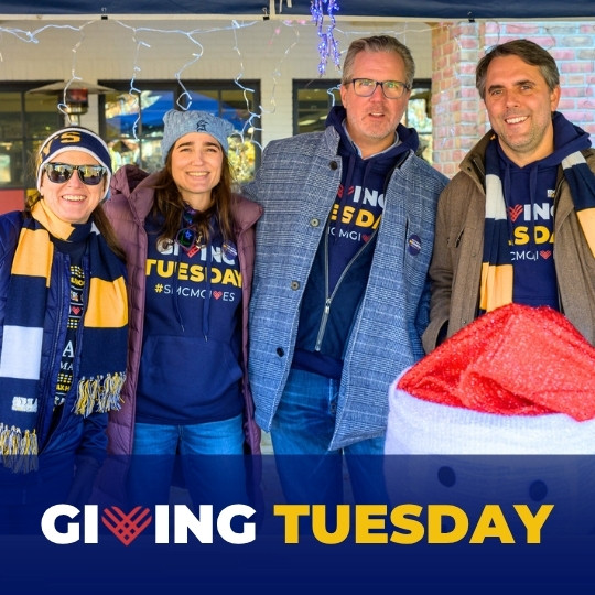 Four people wearing "Giving Tuesday" shirts and scarves stand together and smile at an outdoor event, with a "Giving Tuesday" banner at the bottom of the image.