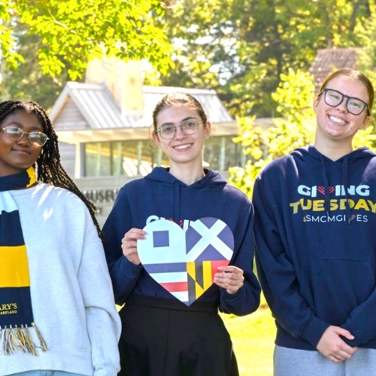 Three students stand outdoors, smiling at the camera. One holds a heart-shaped sign with various designs. They wear sweatshirts and scarves with school colors. Trees and a building are in the background.