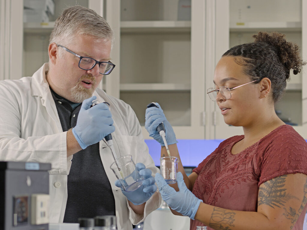 SMCM_AllHonors_LPImage_crop-1024×768 Two scientists wearing gloves and glasses use pipettes to transfer liquid between beakers in a laboratory setting.