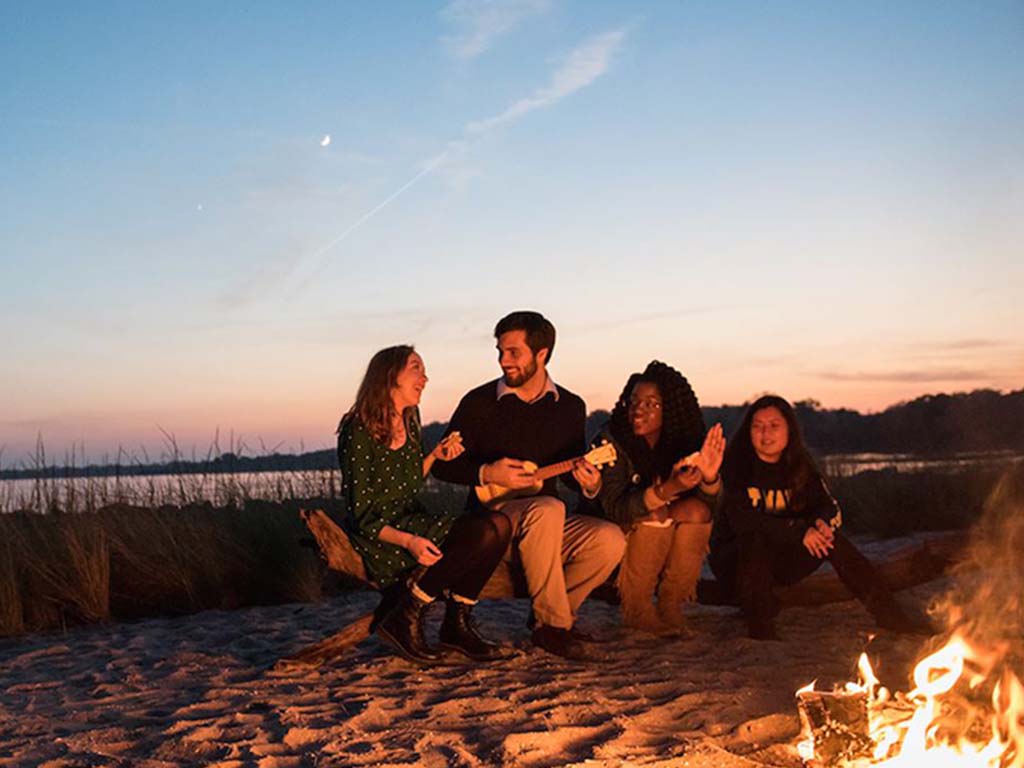 Four people sit by a campfire on a sandy beach at sunset, with one person playing a small guitar. Grassy dunes and water are visible in the background.