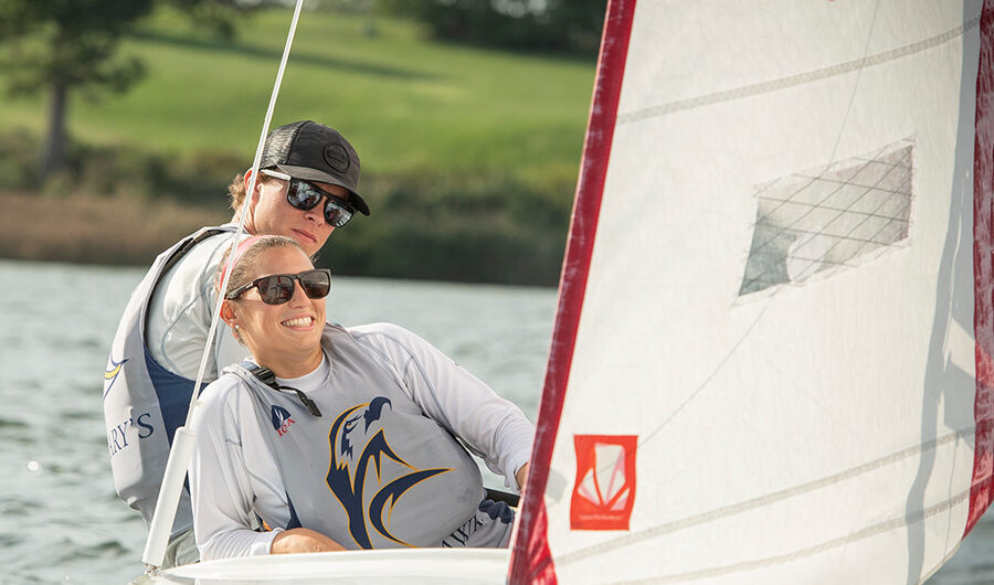 smcm-athletics Two people wearing sunglasses and sailing gear steer a small sailboat on a calm body of water, with grassy land and trees in the background.