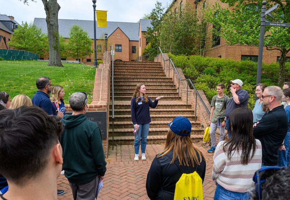 A group of people stands around a tour guide speaking in front of outdoor brick stairs on a college campus.