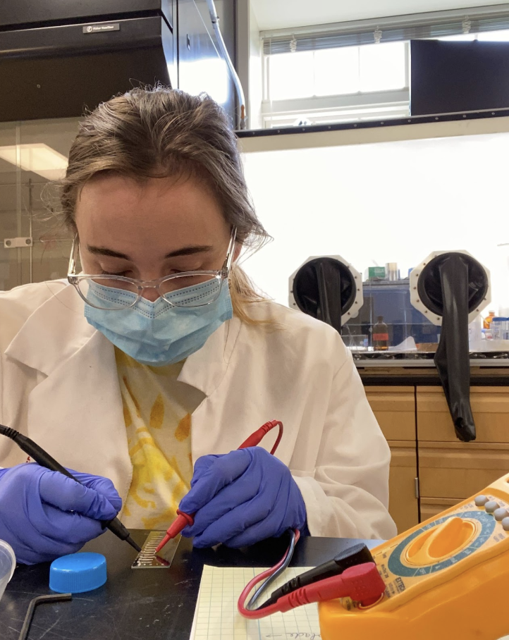 Person in lab coat, mask, and gloves uses probes on a circuit board at a lab workstation; a multimeter and notes are visible.