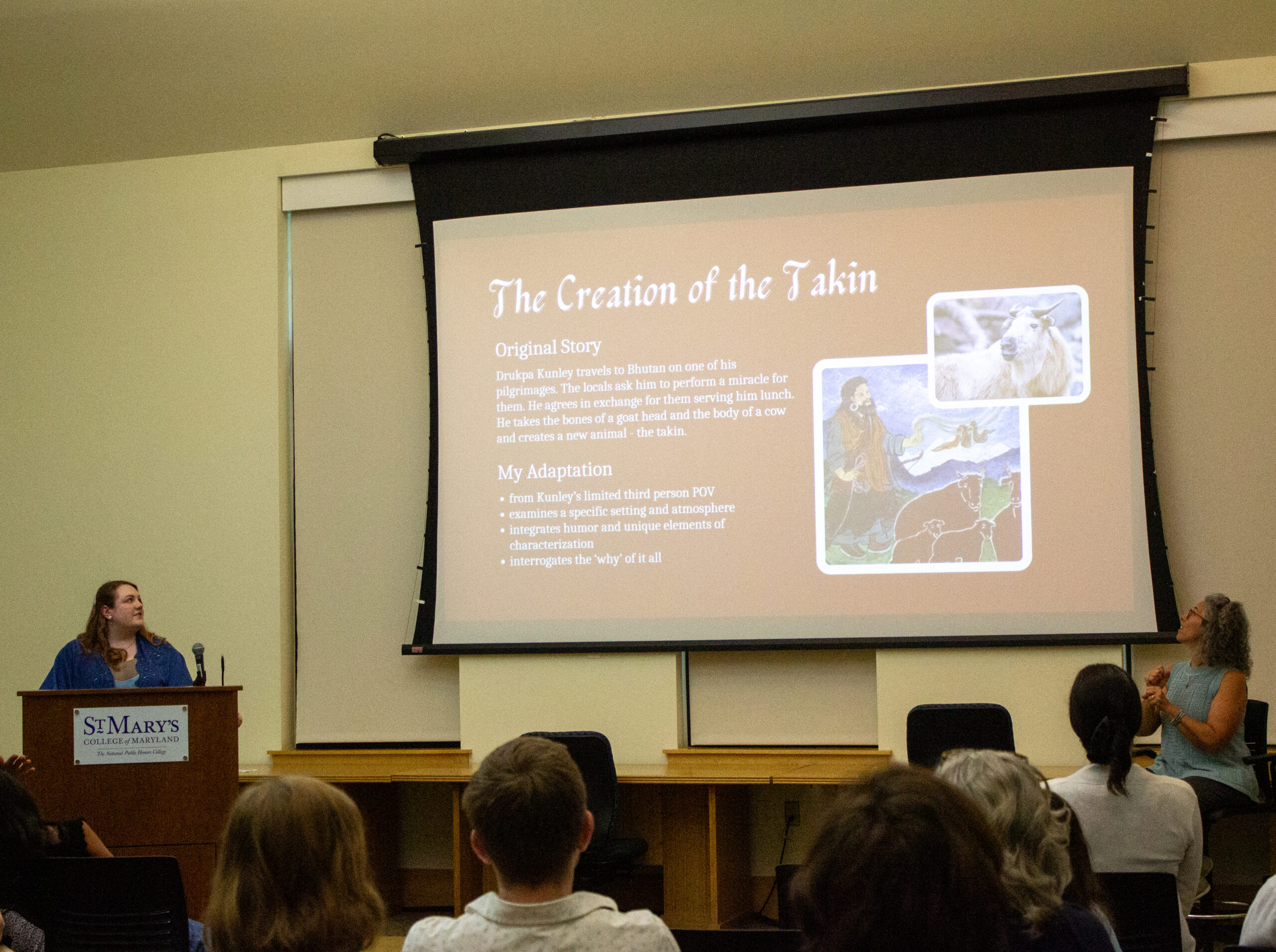 A woman presents a slideshow titled "The Creation of the Jakin" to an audience in a classroom, with text and images displayed on a large screen.