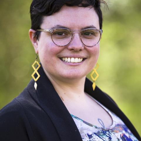 Person with short dark hair, glasses, and geometric yellow earrings smiling at the camera, wearing a black blazer and patterned top, with a blurred green background.