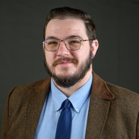 A man with short hair, glasses, and a beard is wearing a brown blazer, blue shirt, and blue tie, standing in front of a plain dark background.