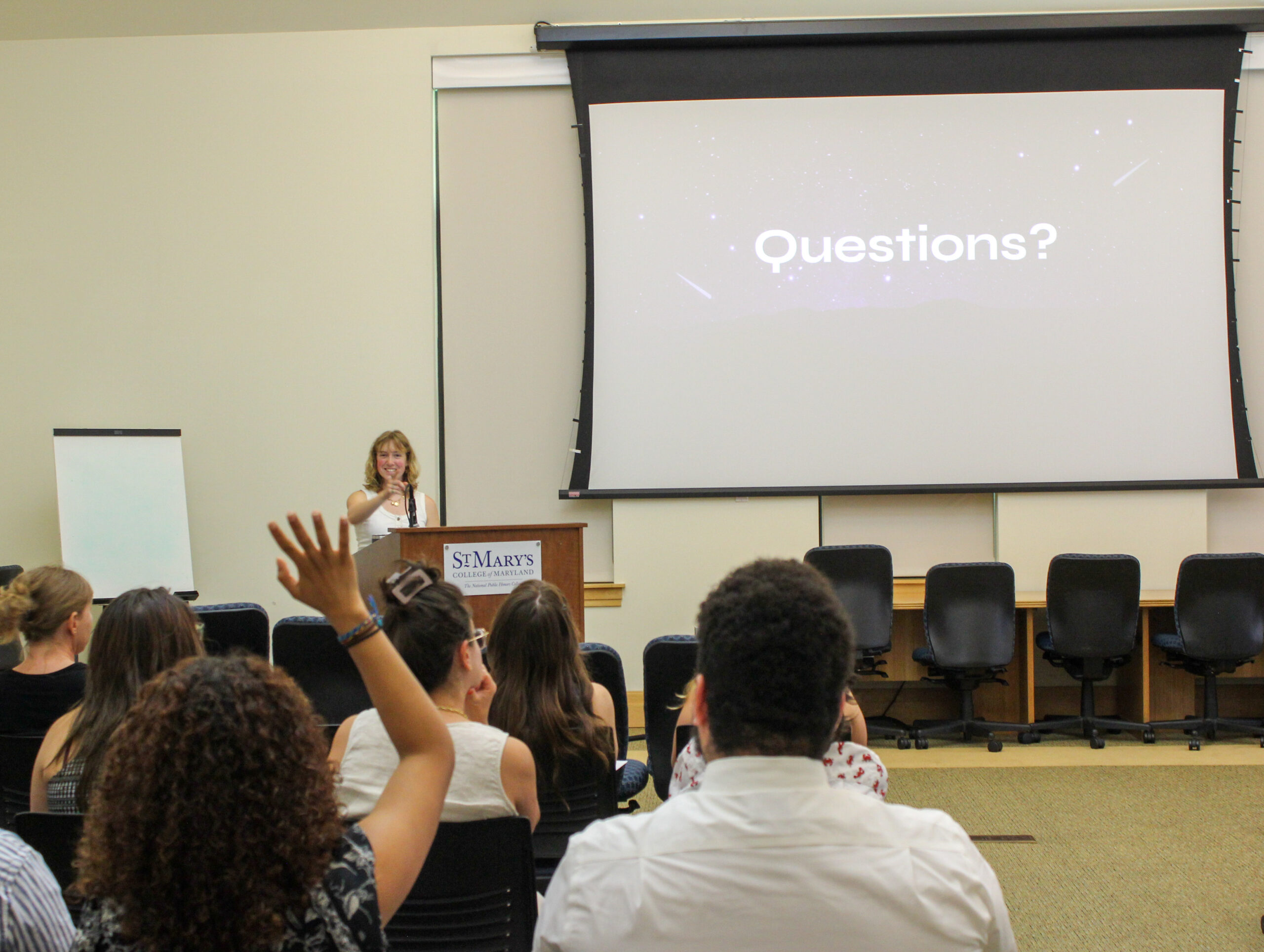 A woman stands at a podium labeled “St. Mary’s” in front of an audience, with a screen behind her displaying the word “Questions?” as someone raises their hand.