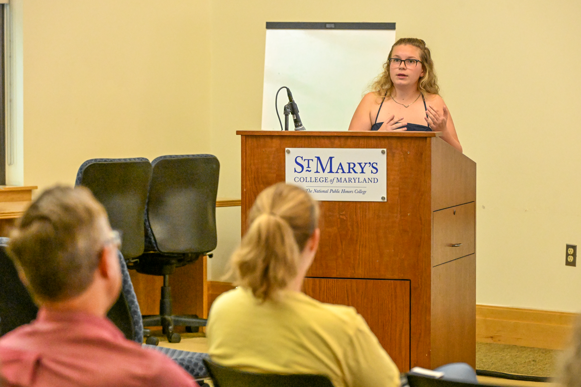 A woman speaks at a podium labeled “St. Mary’s College of Maryland” while two people sit and listen in a classroom setting.