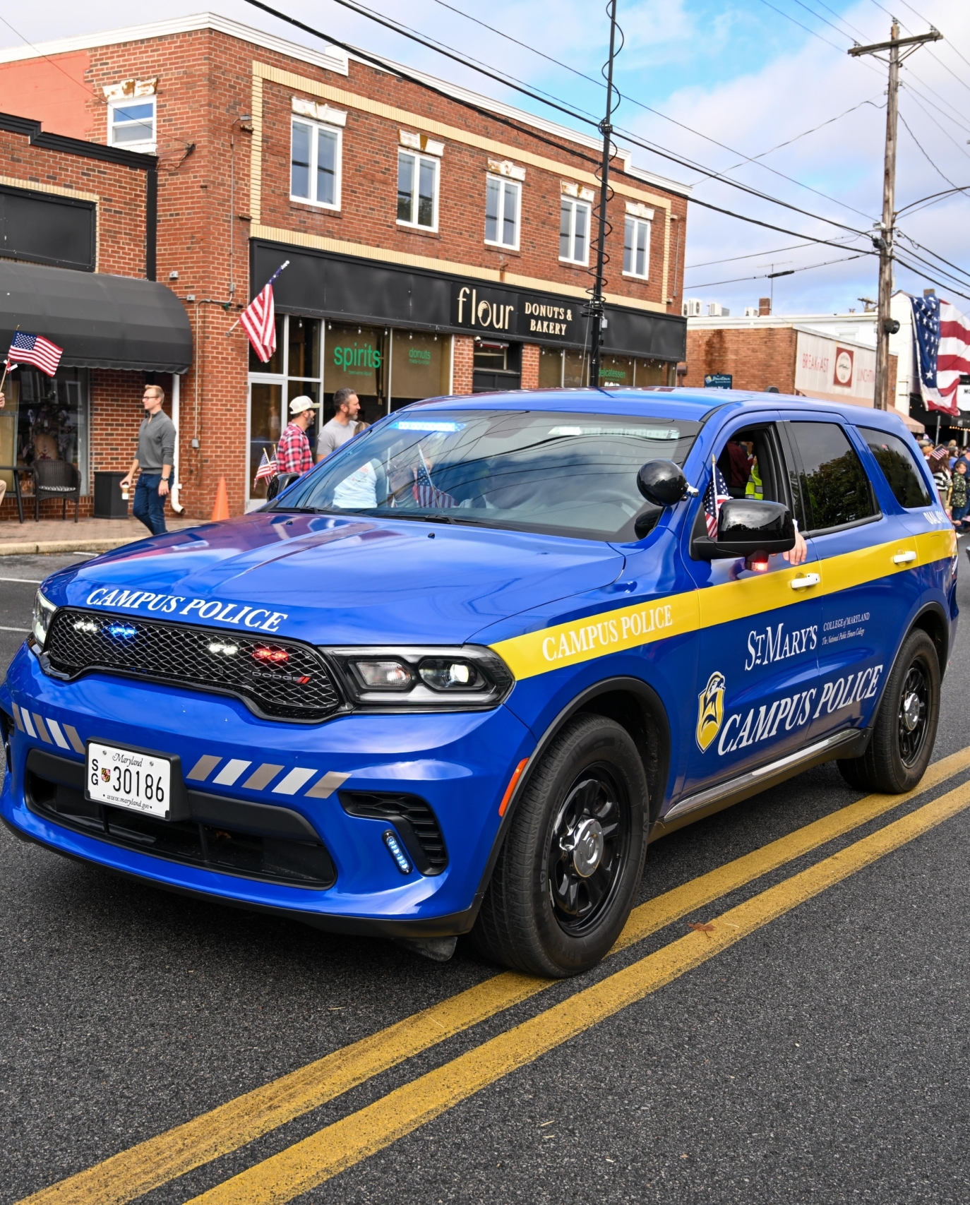 A St. Mary's Campus Police SUV drives down a street during a parade, with people holding American flags on the sidewalk and shops in the background.