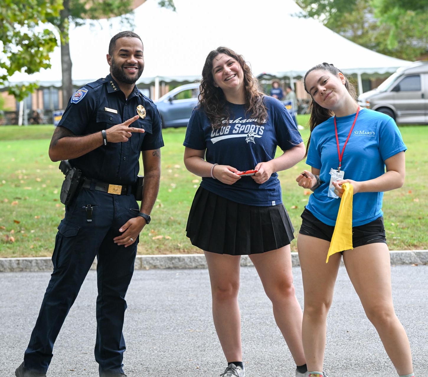A police officer and two young women stand outdoors, smiling at the camera; the women hold papers and a yellow cloth. Trees, vehicles, and tents are visible in the background.