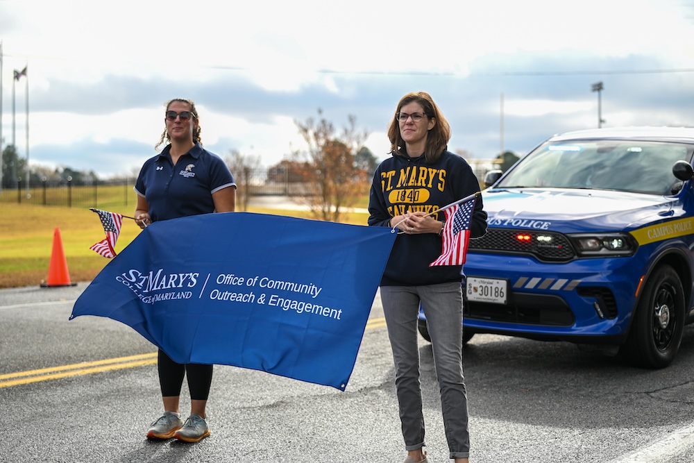 Two women stand on a road holding a St. Mary's College of Maryland outreach banner and small American flags, with a campus police vehicle parked behind them.