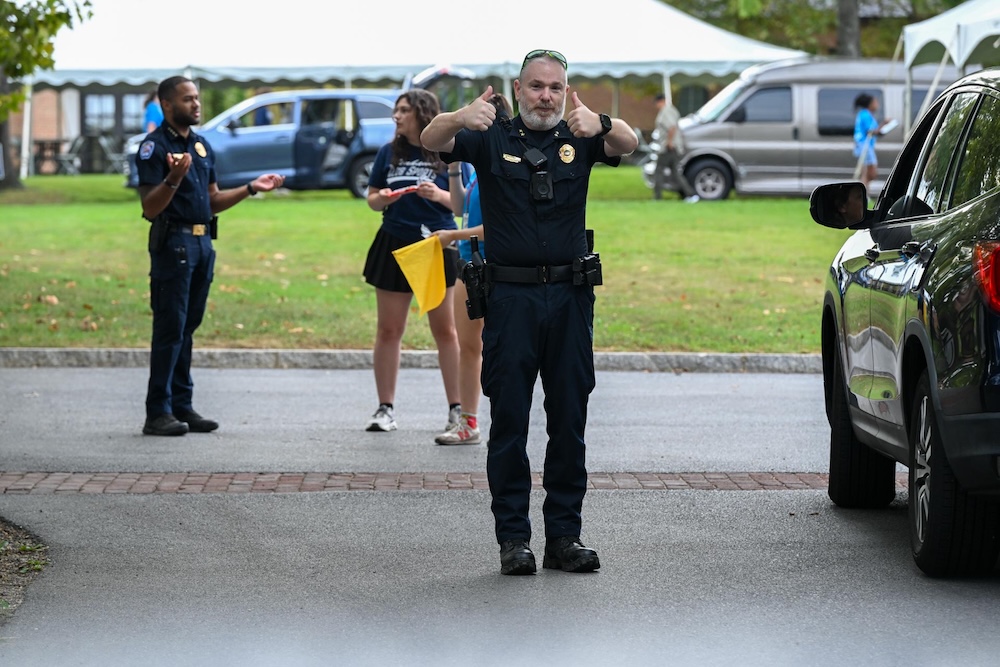 A police officer stands on a road giving two thumbs up, with other people and parked cars visible in the background.