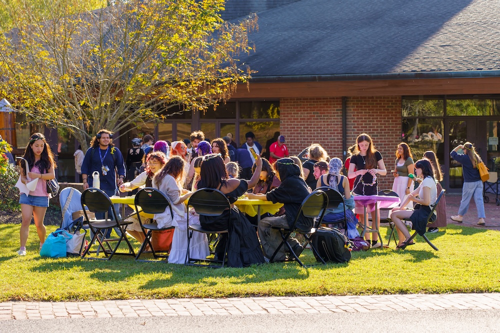 A group of people sit at tables outdoors on a sunny day, with others standing and walking nearby in front of a brick building.