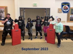 Five people in padded protective gear and one instructor stand in a room with red training pads.