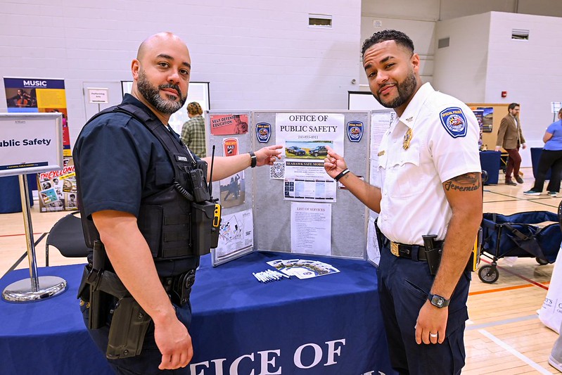 Two public safety officers stand at an informational booth with brochures and a display board labeled "Office of Public Safety" in a gymnasium.