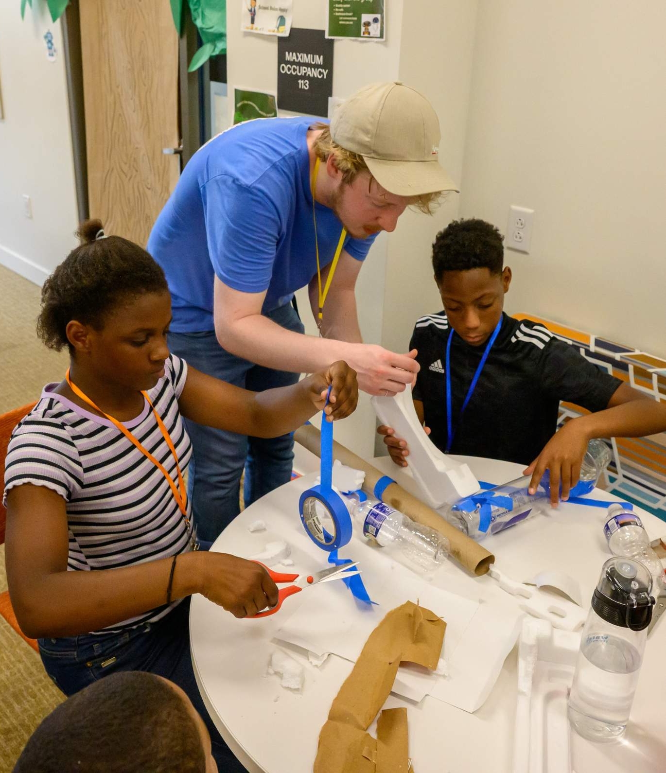An adult assists two children at a table with a hands-on project involving tape, scissors, and recycled materials in a classroom setting.