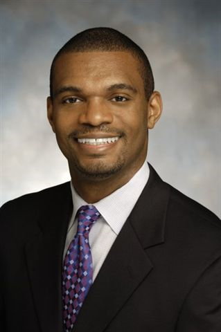 A man in a dark suit, white shirt, and patterned purple tie smiles in a professional studio portrait with a neutral background.
