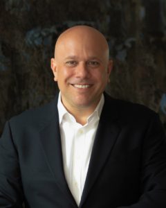 A bald man in a dark suit and white shirt smiles at the camera against a dark, blurred background.