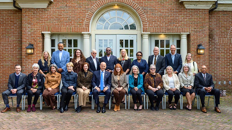 A group of 22 people, dressed in business attire, pose for a formal photo in front of a brick building with large windows and an arched doorway.