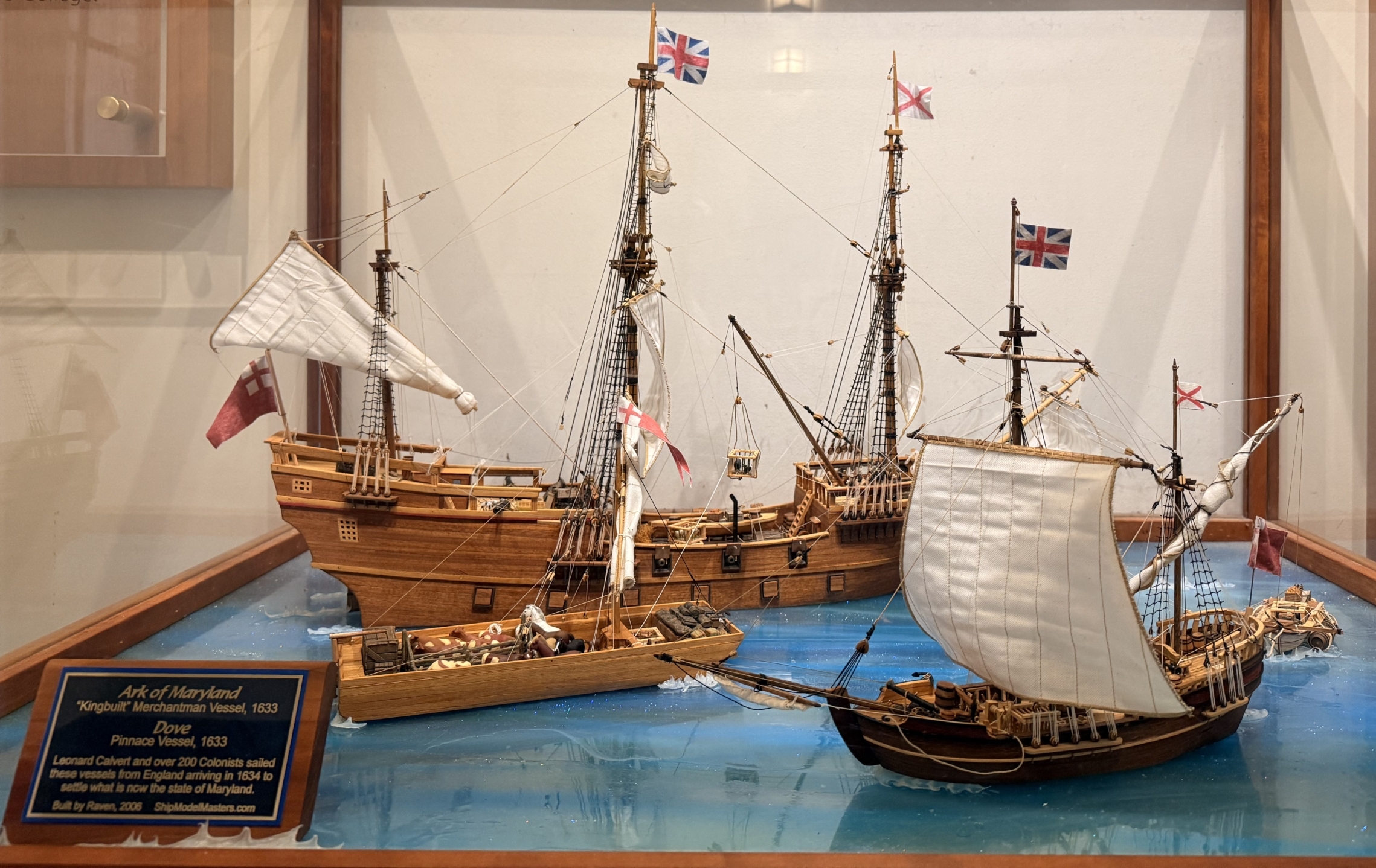 A detailed model display of three historic wooden sailing ships with British flags on a blue water base, encased in a glass box with an informational plaque in front.