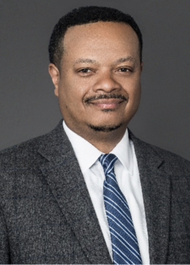 A man in a grey suit, white shirt, and blue striped tie poses for a formal portrait against a plain grey background.