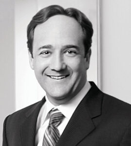 A man in a suit and striped tie smiles at the camera in a professional, indoor setting.