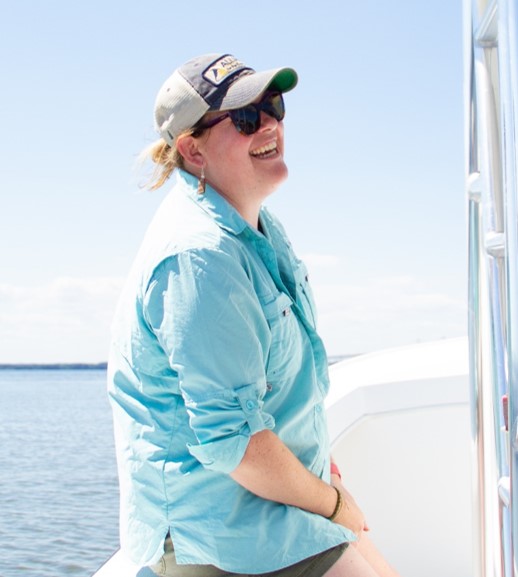 A person wearing sunglasses, a baseball cap, and a light blue shirt smiles while sitting on a boat with water and sky in the background.