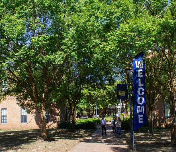 A brick campus building with large trees and a blue "WELCOME" banner; a few people walk along a sunny pathway.