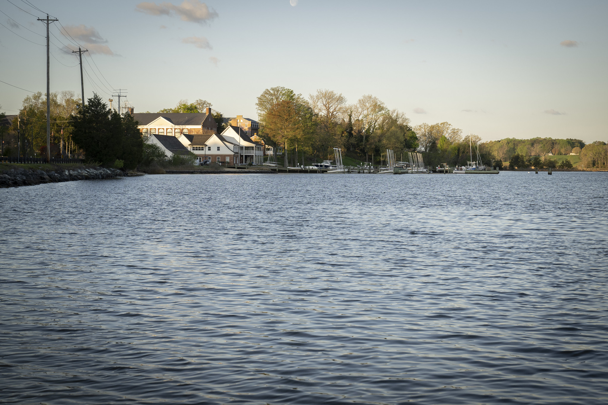 Calm waterfront scene with buildings, trees, and docked sailboats under a clear sky, viewed from across the water.