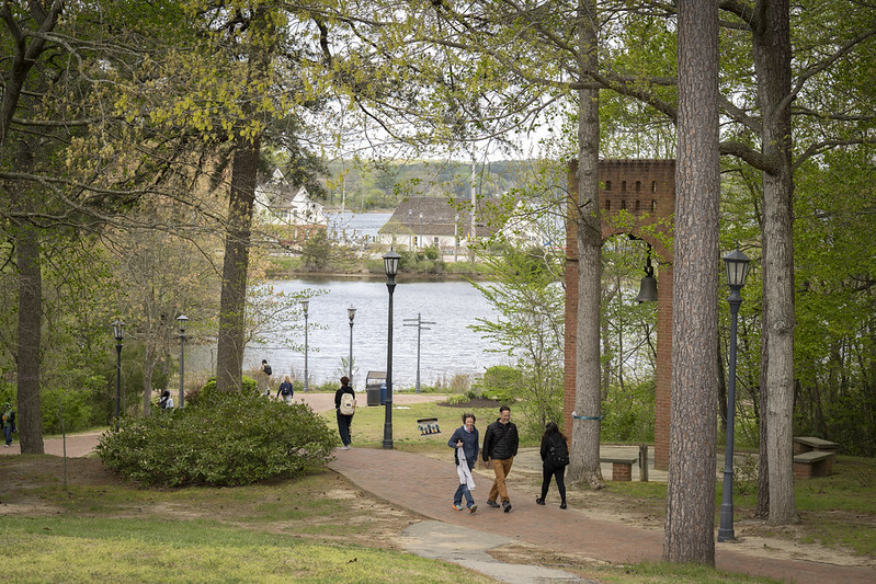 People walk along a paved path in a park with trees, benches, and lamp posts, near a river with buildings visible on the opposite bank.