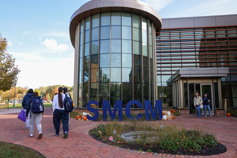 Students walk toward a modern glass building with large blue "SMCM" letters displayed outside on a brick walkway.