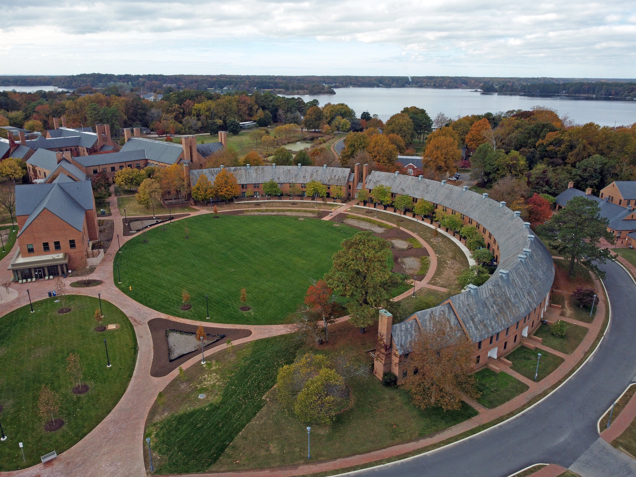 Aerial view of a large, semi-circular brick building surrounding a green lawn, with trees in autumn colors and a lake in the background.