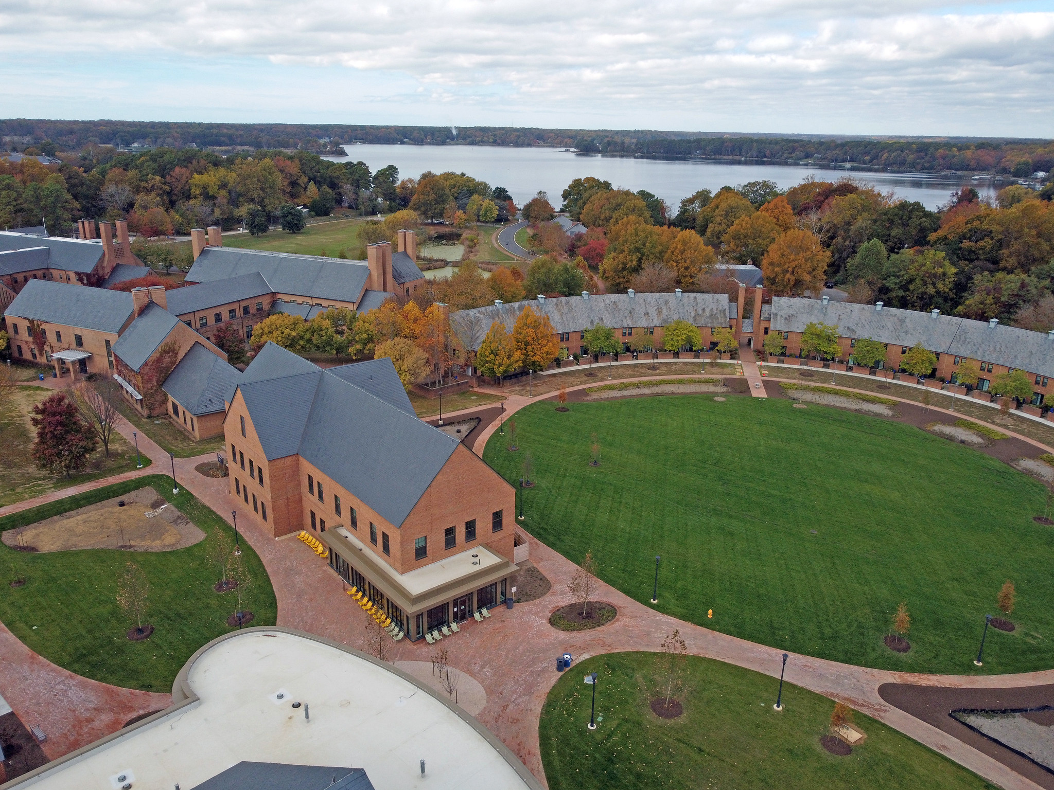 Aerial view of a large, brick academic campus with green lawns, walking paths, and a lake in the background surrounded by autumn trees.