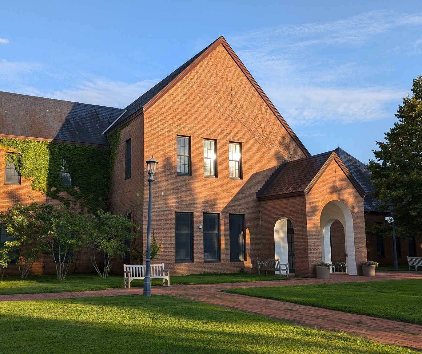 A red brick building with tall windows, an arched entrance, and ivy along one wall, surrounded by green grass, trees, benches, and a brick walkway on a sunny day.
