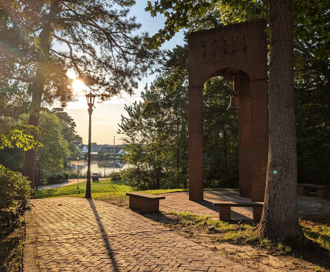 A brick archway with a bell stands near a paved path and benches, surrounded by trees, with sunlight filtering through and a river visible in the background.