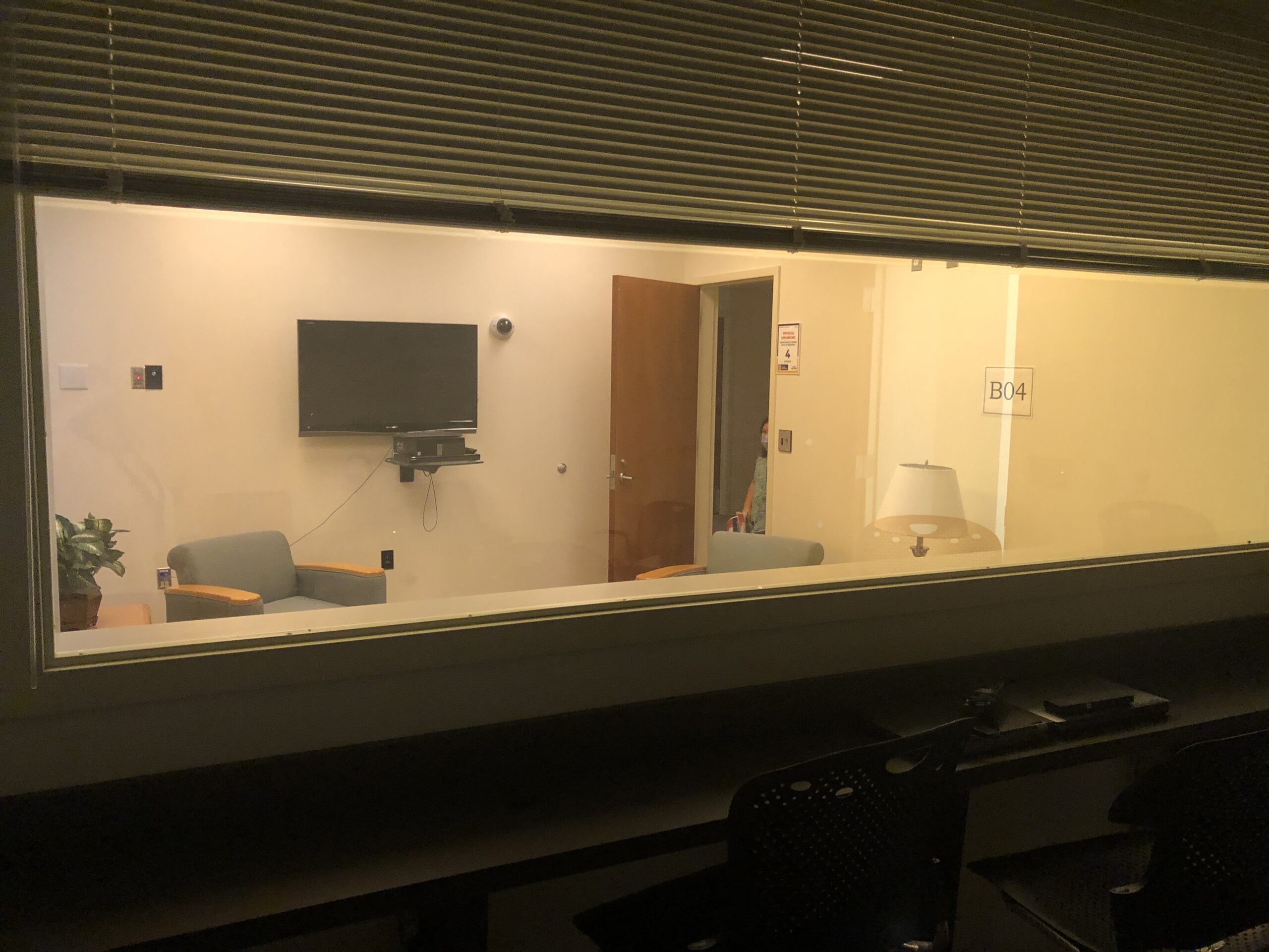 A view through a window into a small office with two chairs, a TV on the wall, a plant, a door, and a sign labeled “B04.”.