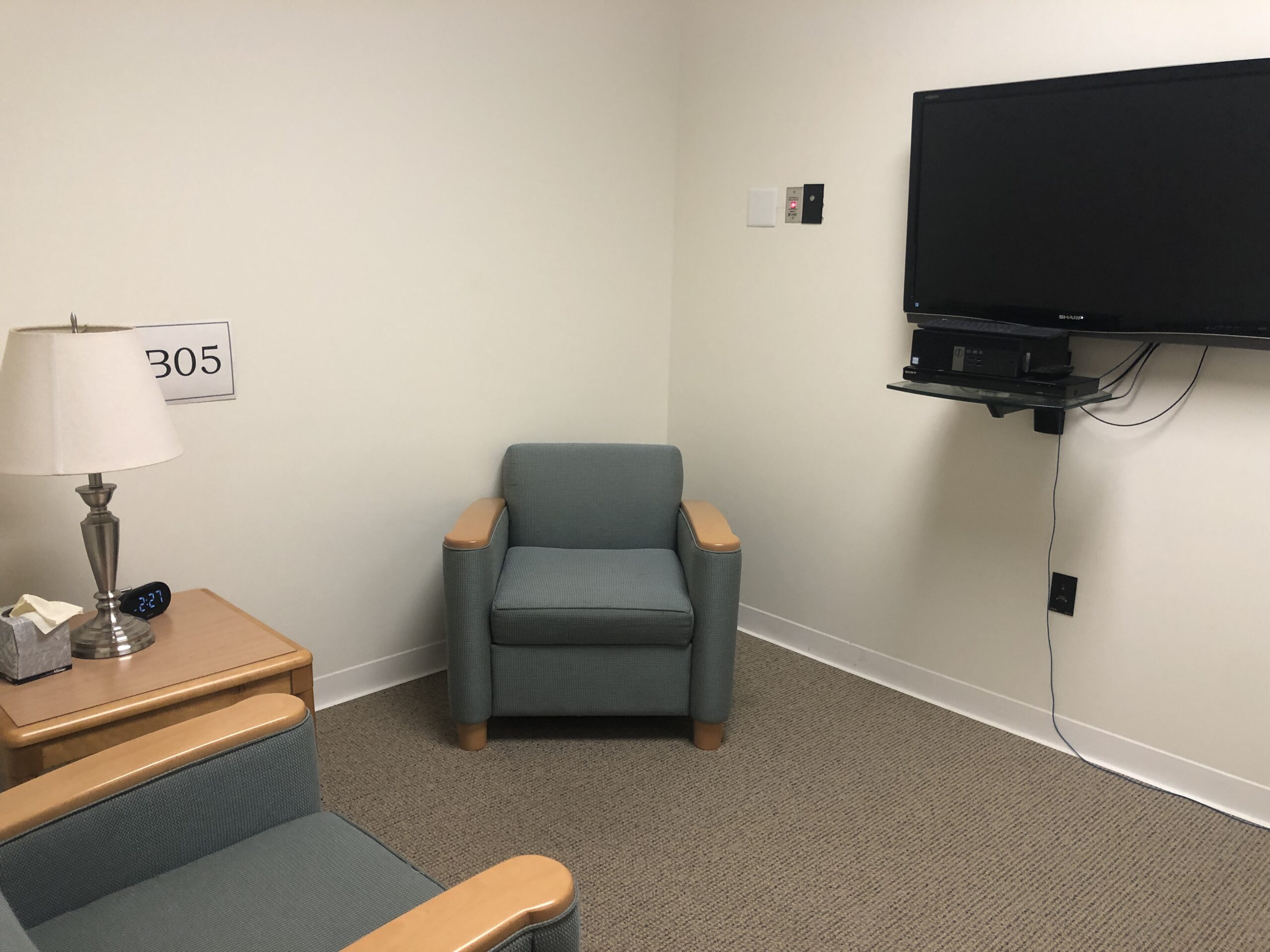Small office with two blue armchairs, a lamp, tissue box, wall-mounted TV, and a digital clock on a side table. Room number 305 is on the wall.
