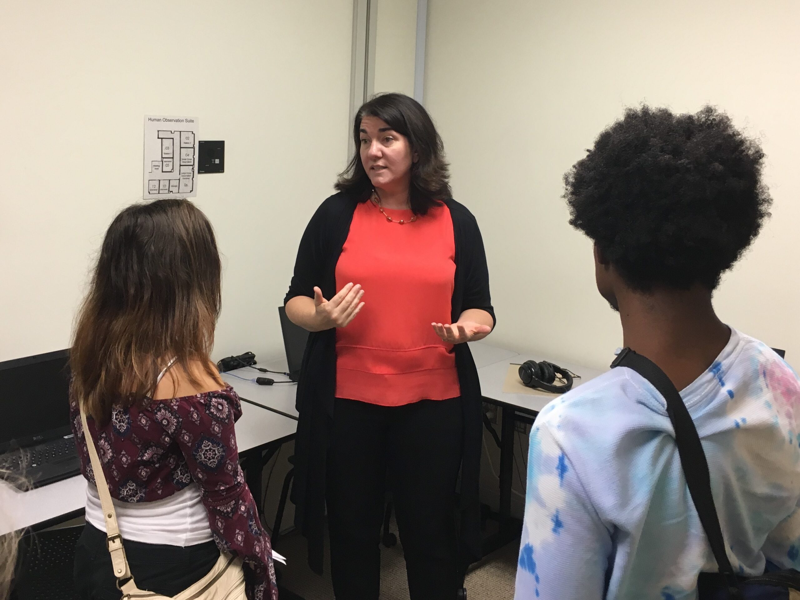 A woman stands and speaks to three students in a classroom with computers and headphones visible in the background.