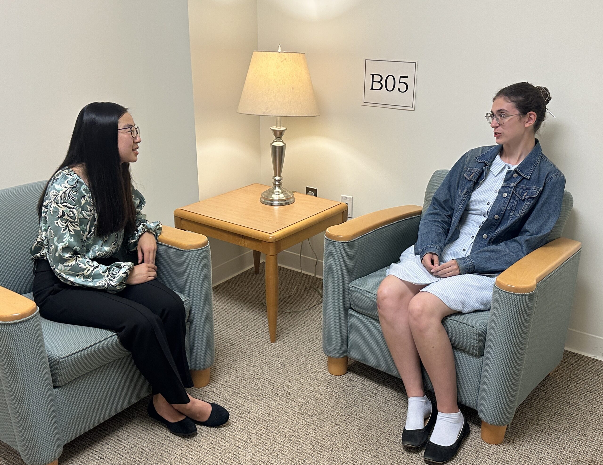 Two women sit facing each other in armchairs by a side table with a lamp in a room labeled “B05.”.