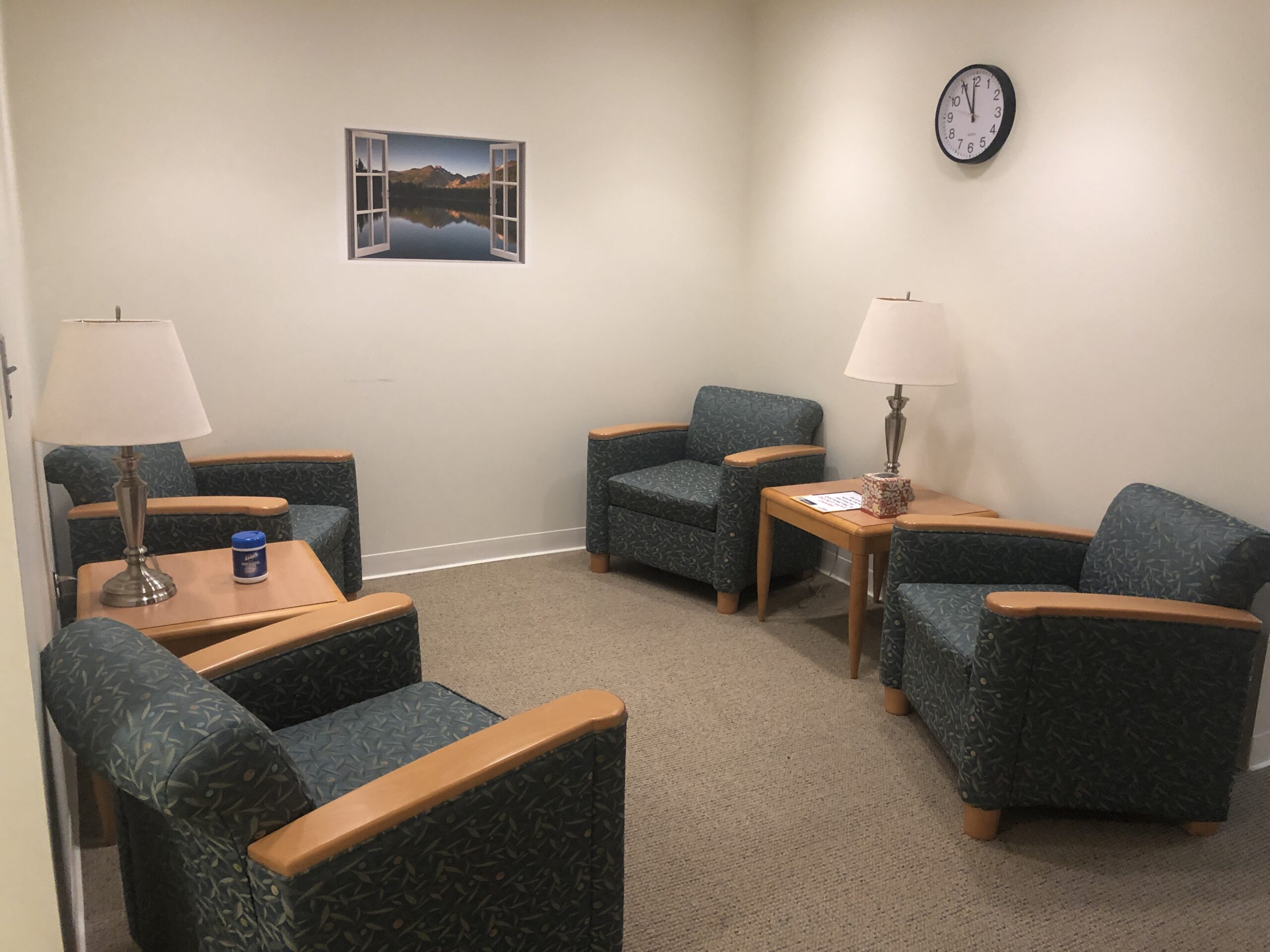 A small waiting room with four green armchairs, two side tables, two lamps, a clock on the wall, and a window painting above one chair.