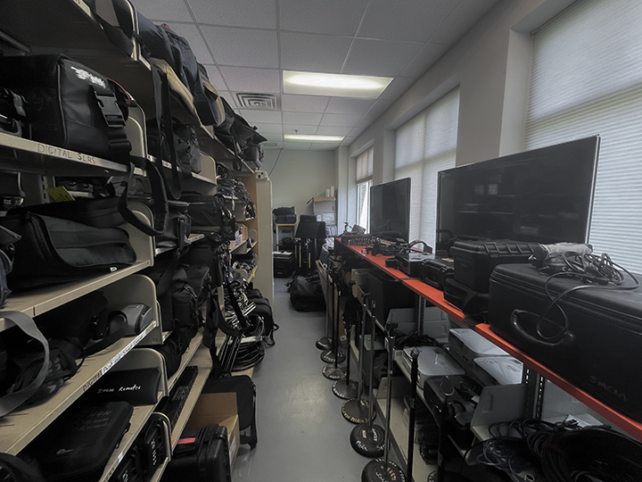 A storage room with organized shelves holding camera bags, cases, tripods, monitors, and various technical equipment along both walls.