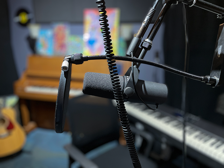 A close-up of a microphone with a pop filter in a recording studio, with a piano, keyboard, and acoustic guitar visible in the blurred background.