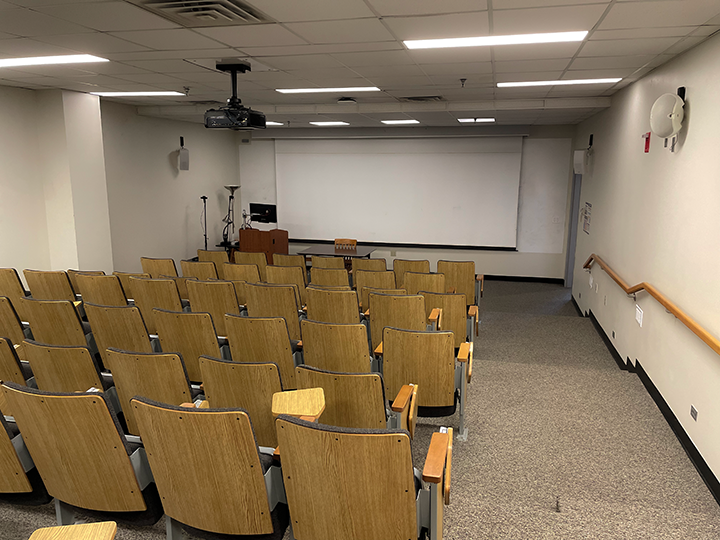 Lecture hall with rows of empty wooden chairs, a projector on the ceiling, a podium, and a large blank screen at the front.