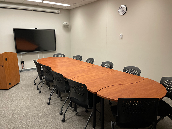 A conference room with a long wooden table, ten black chairs, a wall-mounted screen, a podium, clock, and neutral-colored walls.