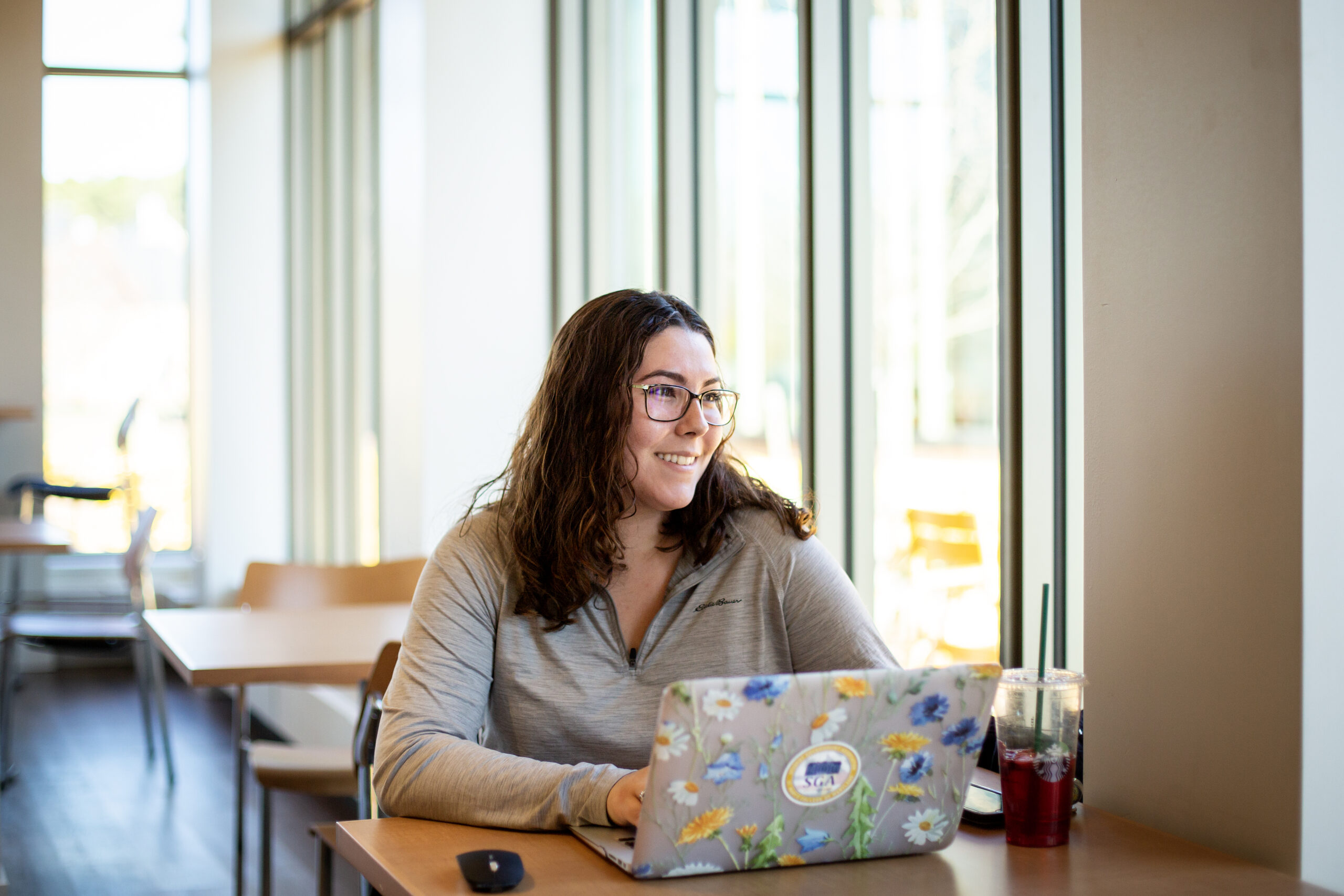 A woman with glasses sits at a table by large windows, working on a laptop decorated with flower stickers. A drink with a straw is next to her on the table.