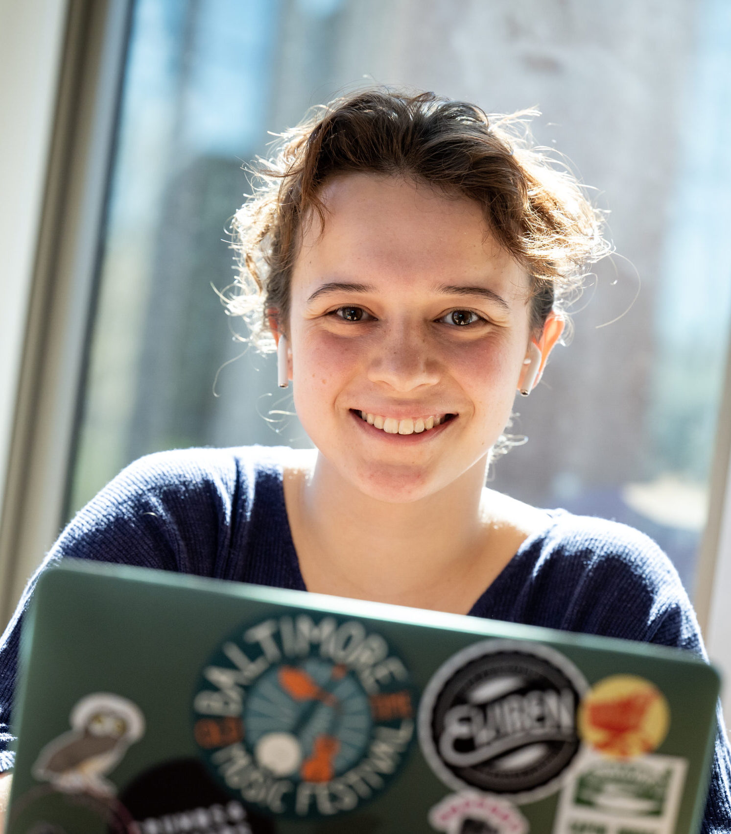 A person smiling at a table, working on a laptop decorated with various stickers, with sunlight coming through a window behind them.