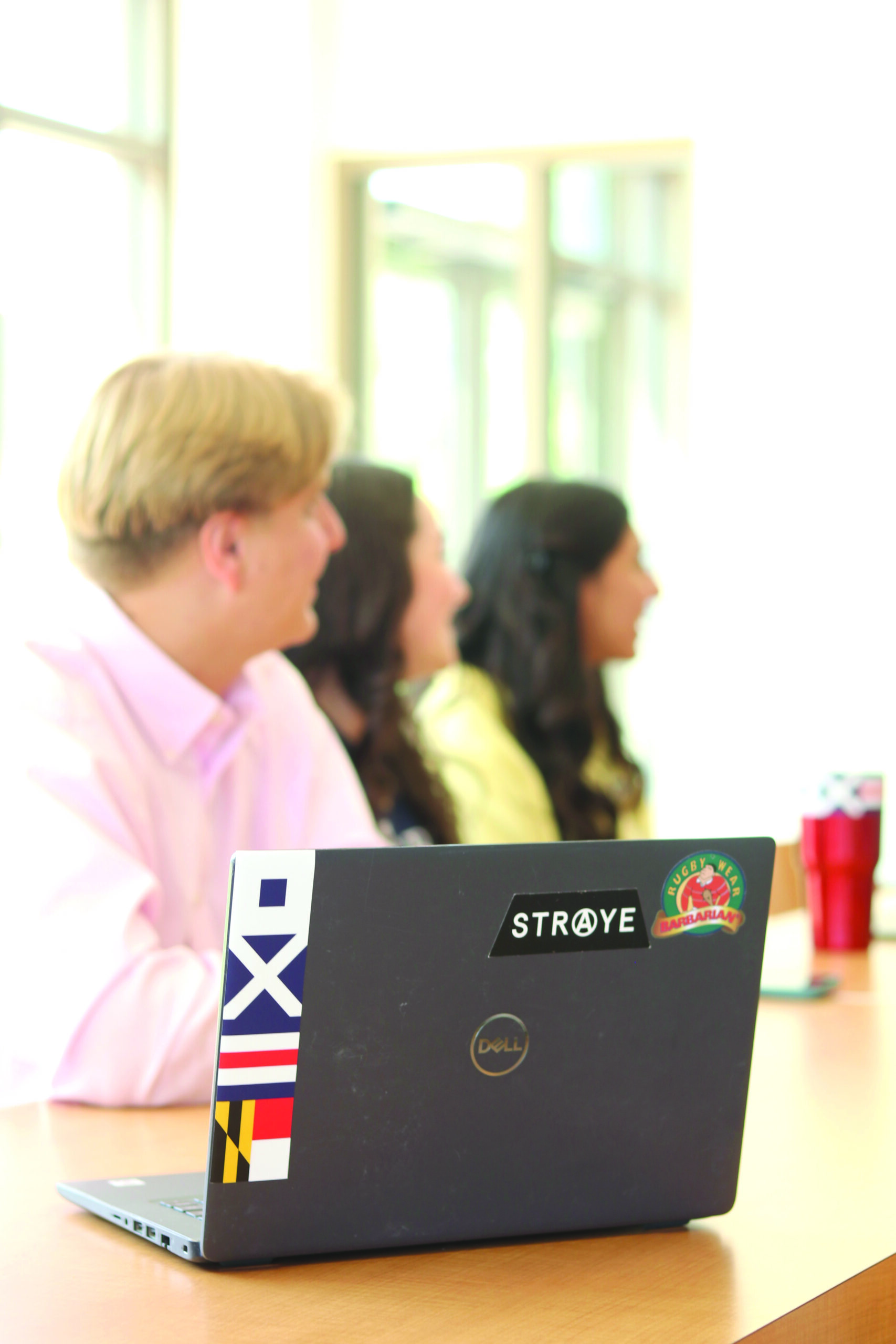 Three people seated at a table are out of focus, while a Dell laptop with several stickers is in clear view in the foreground.