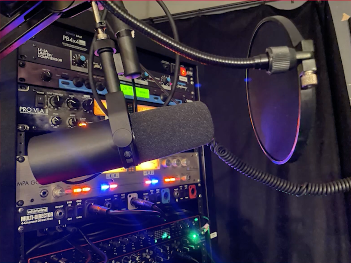 Close-up of a professional recording setup with a microphone, pop filter, and audio equipment rack featuring various controls and illuminated displays.